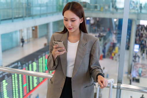 Young asian woman carrying suitcase and checking smartphone on hand in airport terminal. Ready to travel. Travel and vacation concept. Business person on business trip. Wireless on the move. Digital banking anytime, anywhere