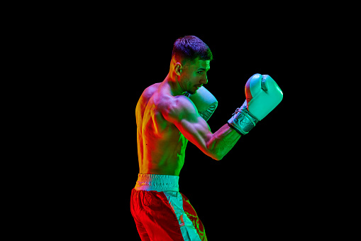 Side view portrait of sportsman boxer, mixed martial art fighter workout against black mode background in mixed neon filter, light. Concept of sport, action, motion, healthy lifestyle. Copy space, ad.