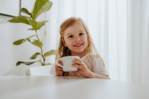 A smiling girl having a cup of milk while sitting at the kitchen desk and looking at camera.