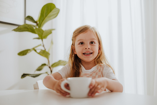 Cheerful girl having a cup of milk while sitting at the kitchen desk and looking at camera.