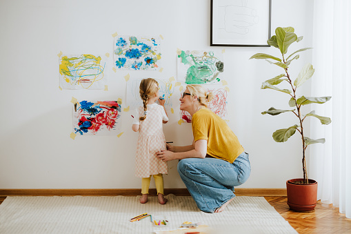 A smiling mom watching how her unrecognizable daughter painting with a felt tip pen on a paper hanging on a nursery wall.
