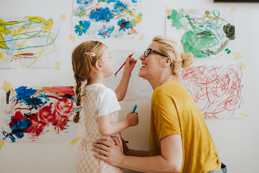 Happy mom and daughter laughing and painting with a felt tip pens together on a white paper hanging on the living room wall at home.