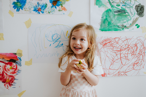 Cheerful smiling young girl holding a fresh apple while standing in front of a white wall with her drawings and laughing.