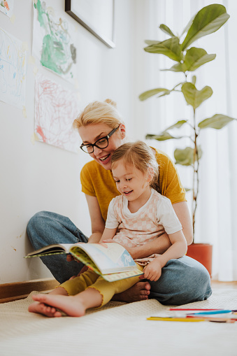 Smiling mother embracing her little girl while sitting on the floor with a book, laughing and reading the story.