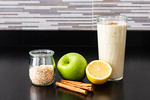 A glass with homemade smoothie next to half lemon, one green apple, a glass pot with oat flakes and two cinnamon sticks over a black kitchen bench.