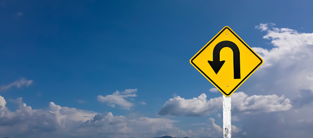 Traffic sign: Left U-turn sign on cement pole beside the rural road with white cloudy bluesky background, copy space.