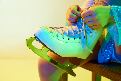 Close-up image of skates. Girl, child tying laces on skates before dancing against yellow background in neon light. Concept of childhood, figure skating sport, hobby, school, education