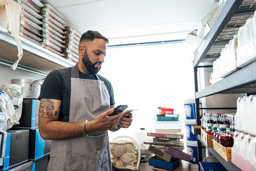 Side view of a mid adult Indian man wearing all black casual clothing and an apron. He is working in the back of his family-run fish and chip shop in Gateshead, England. He is performing a stock check using his smartphone.