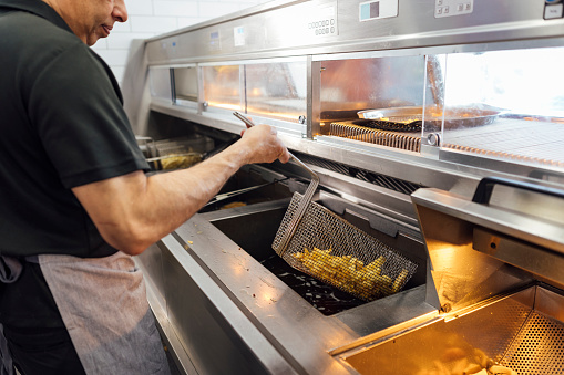 Side view of a mature Indian man wearing all black casual clothing and an apron. He is working in his family-run fish and chip shop in Gateshead, England. He uses a deep-fryer to fry chips.

Videos also available for this scenario.