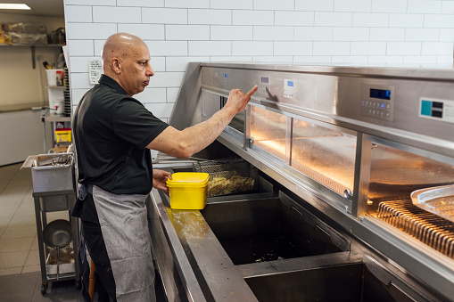 Side view of a mature Indian man wearing all black casual clothing and an apron. He is working in his family-run fish and chip shop in Gateshead, England. He uses a deep-fryer to fry fish and chips.

Videos also available for this scenario.
