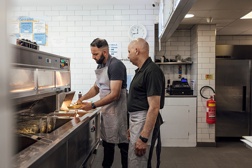 Three quarter length side view of a mature Indian man and his mid adult son wearing all black casual clothing and aprons. They are working in their family-run fish and chip shop in Gateshead, England. The mid adult man is serving a portion of kebab meat and chips with garlic sauce.
