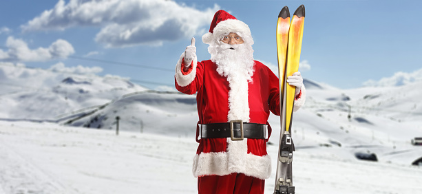 Santa Claus at a mountain holding a pair of skis and gesturing thumbs up