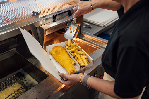 An over the shoulder view of an unrecognisable mature Indian woman wearing all black casual clothing and an apron. She is working in her family-run fish and chip shop in Gateshead, England. She is scooping fish and chips into a box ready to serve.\n\nVideos also available for this scenario.