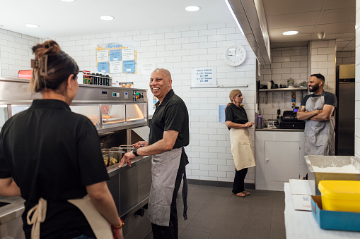 Side view of a mature Indian couple, their mid adult son and his wife wearing all black casual clothing and aprons. They stand together in their family-run fish and chips shop, chatting and working in Gateshead, England.