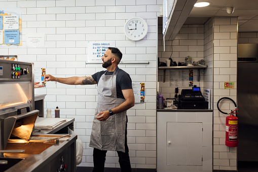 Side view of a mid adult Indian man wearing all black casual clothing and an apron is serving an unrecognisable customer passing over their change, he is working in a family-run fish and chip shop in Gateshead, England.