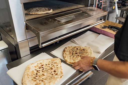 Close up view of an unrecognisable mature Indian woman working in her family-run fish and chip shop in Gateshead, England. She is placing a flatbread in the oven to cook.