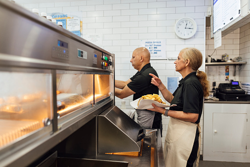 Three quarter length side view of a mature Indian man and his mature wife wearing all black casual clothing and aprons. They are working in their family-run fish and chip shop in Gateshead, England. The mid woman is serving a portion of kebab meat and chips in a wrap.