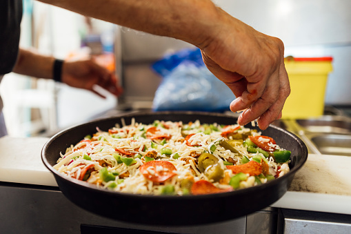 Close up view of an unrecognisable mature Indian male chef making a pizza and sprinkling grated cheese on top before cooking in a pizza oven, he is working in a family-run fish and chip shop in Gateshead, England. Jalapeños, green bell peppers, chicken, red onion and pepperoni are the other pizza toppings.