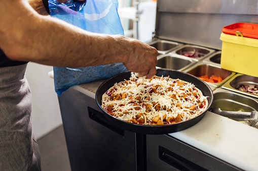 Close up side view of an unrecognisable mature Indian male chef wearing all black casual clothing and an apron making a chicken pizza and sprinkling grated cheese on top before cooking in a pizza oven, he is working in a family-run fish and chip shop in Gateshead, England.