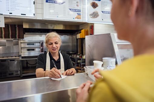 Over the shoulder view of an unrecognisable woman ordering food at the counter of a chip shop. A mature Indian woman wearing all black casual clothing and an apron is serving the customer, she is working in a family-run fish and chip shop in Gateshead, England. She is writing down the customers order with a note pad and pen.

Videos also available for this scenario.