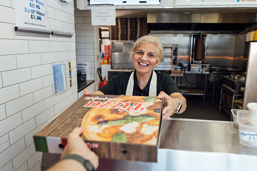 Customers point of view of a mature Indian woman wearing all black casual clothing and an apron serving the customer and passing over a pizza box, she is working in a family-run fish and chip shop in Gateshead, England. She is looking at the customer with a smile.

Videos also available for this scenario.