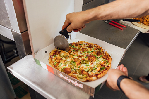 Close up side view of an unrecognisable mature Indian man preparing a pizza box with a freshly made chicken pizza inside, he is cutting the pizza with a pizza cutter working in a family-run fish and chip shop in Gateshead, England.