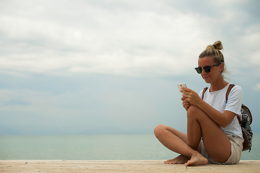 Young tourist woman is sitting on a pier and using mobile phone