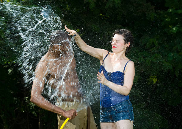 Fun slice of summertime life image. A brunette smashes a water balloon over her friends head as he tries to ward her off with a garden hose.