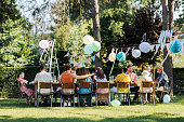 Family and friends sitting at the party table during a summer garden party outdoors. Rear view of people sitting at the table.