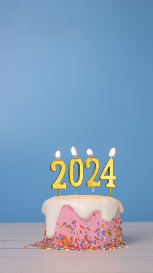 Happy New Year 2024, pink birthday cake decorate with colorful sugar sprinkle and whip cream with golden candle 2024 for new year celebrate party with copy space isolated on blue background, vertical