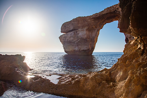 The Azure Window in sunset, Gozo island. The arch is located on the edge of a cliff overlooking the ocean. The arch is made of orange-brown rock. The sea is a deep blue and is visible through the arch. The sky is a clear blue and the sun is shining, creating a warm glow on the rock. The background consists of a rocky coastline and the horizon.