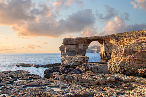 The Azure Window in susnset on Gozo island - a natural rock formation on the coast of a body of water. The rock formation is a natural arch with a flat top. The rock formation is made of a light colored stone. The water is a deep blue and the sky is a light blue with white clouds. The sun is setting, casting a warm glow on the scene. The foreground consists of rocky terrain with small pools of water.