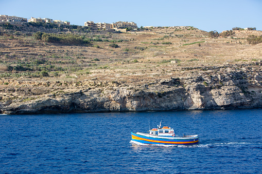 A colorful boat with a blue and orange striped hull is sailing on the Mediterranean Sea. In the background, there is a cliffside village with white buildings and terraced fields. The sky is clear and blue. The photo is taken from a distance, with the boat in the lower right corner and the village in the upper left corner.