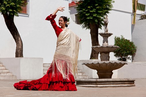 Woman traditional Spanish Flamenco dancer dancing in a red dress and cream shawl dancing in a town square with a stone fountain