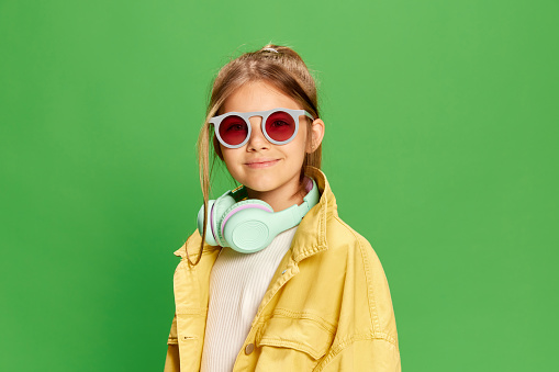 Portrait of little girl wearing stylish clothes and sun glasses over green background with smile looking at camera. Concept of technology, new generation, childhood, lifestyle, ad.