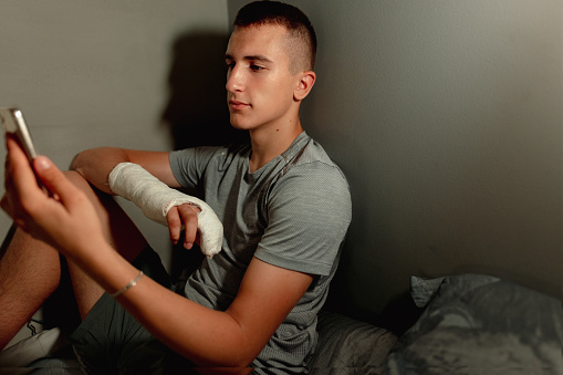 Teenager sitting on his bed using his phone in a dimly lit room with a cast on his right arm