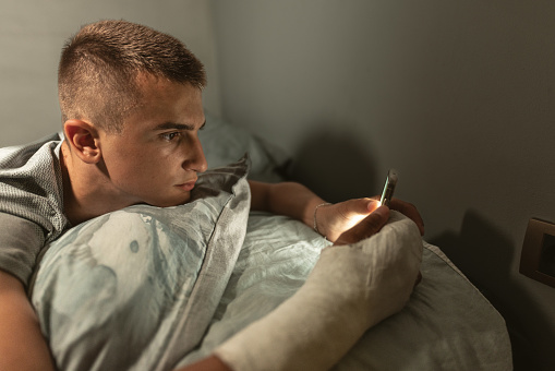 Teenager lying in bed on his phone in a dark room with a cast on his right arm