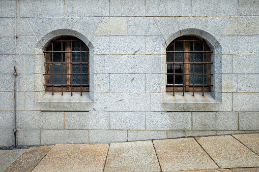 Two small wooden arched windows protected by grilles. The windows set into a solid wall built of granite blocks, in front of which you can see part of the stone slab pavement.