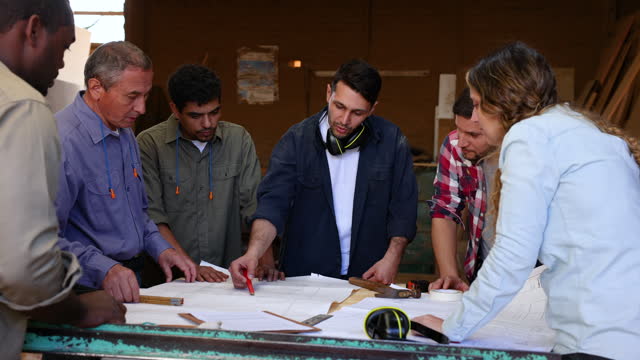 Focused team of people paying attention to male supervisor while he is pointing at a blueprint and explaining something at a manufacturing furniture factory