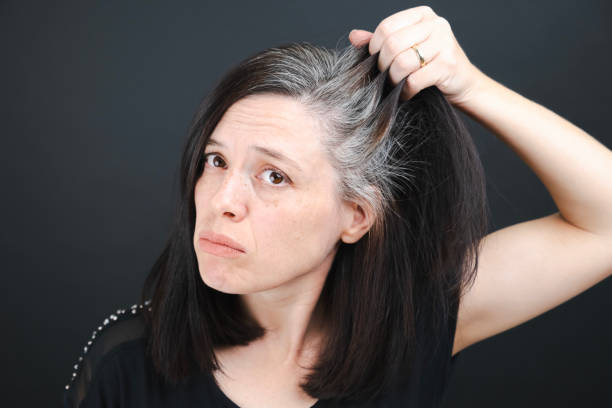 une jeune femme examine les cheveux gris sur sa tête dans un miroir sur fond noir. gros plan sur la texture des cheveux gris. - cheveux blancs photos et images de collection