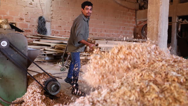 Male worker at a lumberyard moving sawdust with a wooden shovel to a pile
