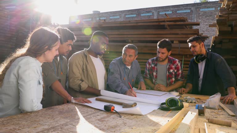 Group of diverse employees in a meeting at a wood factory looking at a furniture design on blueprint while talking