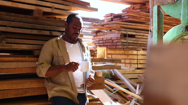 Tired African American worker relaxing on his coffee break at a lumberyard leaning against a pile of wood