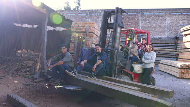 Group of diverse employees working at a lumberyard and sitting outdoors next to a forklift while looking at the camera smiling