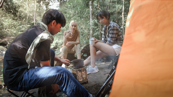 Young People Trekking Group Sitting Relaxed in the Rainforest and Boil Water with a Camp Stove. Campsite Drinking Coffee and Water. Prepare Easy Camping Meals for Groups on Trekking Trips.