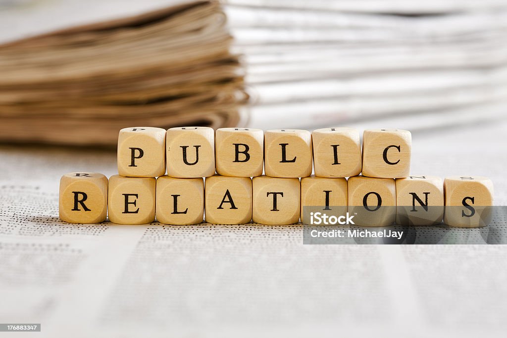 Letter Dices Concept: Public Relations Concept of dices with letters forming words: Public Relations. Generic newspaper background with some blurred text on the bottom and paper stack in the back. Abstract Stock Photo