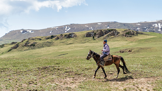 Kyrgyzstan - May 2022: Kyrgyz man on horseback riding in Kyrgyzstan mountains. In Kyrgyzstan's mountainous regions, it is common to see shepherds tending to their flocks while riding on horseback.