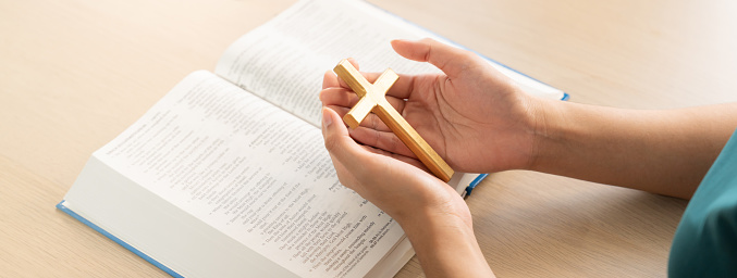 Female god believer holding wooden cross on opened holy bible book at light wooden church table. Top view. Concept of hope, religion, faith, christianity and god blessing. Warm background. Burgeoning.