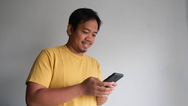 mężczyzna z azji południowo-wschodniej uśmiecha się i używa telefonu komórkowego na szarym tle - southeast asian ethnicity men laptop image type zdjęcia i obrazy z banku zdjęć