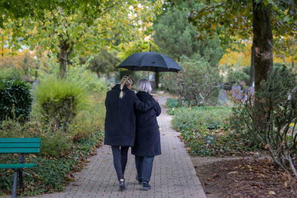 Rear view of daughter holding her mother on the way to the man's grave Rear view of daughter holding her mother in her arms on the way to the man's grave in the cemetery on a cloudy cold day annoying mother in law stock pictures, royalty-free photos & images